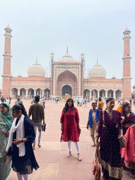 Transcend's founder, Nazia is standing in front of the historic Jama Masjid in old Delhi, India. She is wearing a red linen kurta and white pants