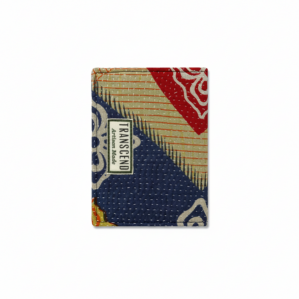 A square fabric accessory, likely a wallet or small pouch, featuring a colorful patchwork kantha quilt pattern with stitched blue, red, beige, and white sections. A white label on the front reads 'TRANSCEND Artisan Made'.