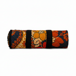 A rolled-up quilted yoga mat featuring a vibrant floral and paisley pattern in orange, red, navy blue, and beige, secured with black straps on a white background. 1