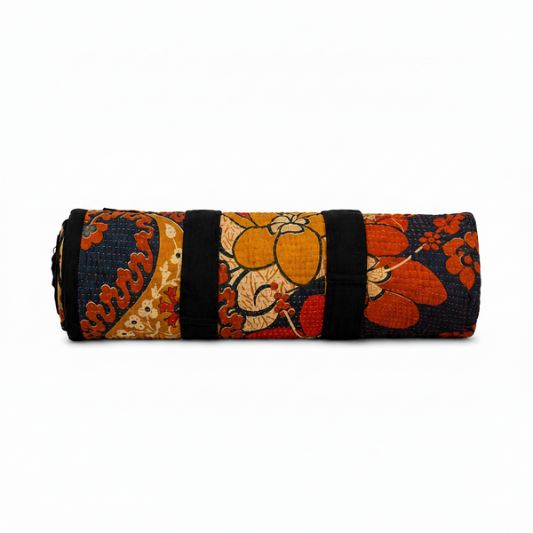 A rolled-up quilted yoga mat featuring a vibrant floral and paisley pattern in orange, red, navy blue, and beige, secured with black straps on a white background.