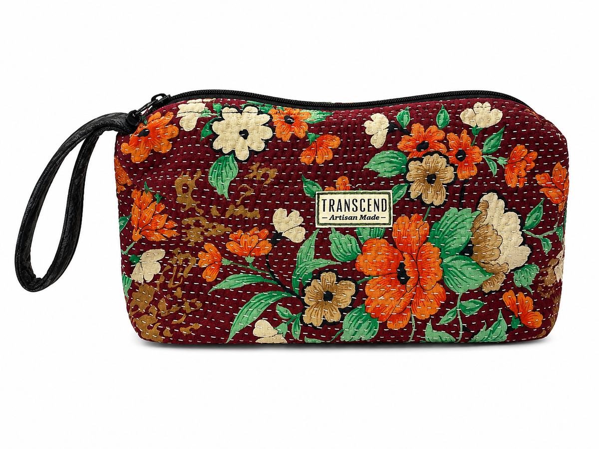 A rectangular zippered pouch with a wrist strap, featuring a vintage floral pattern in orange, beige, and green on a dark red background. A small white label on the front reads 'TRANSCEND Artisan Made'.