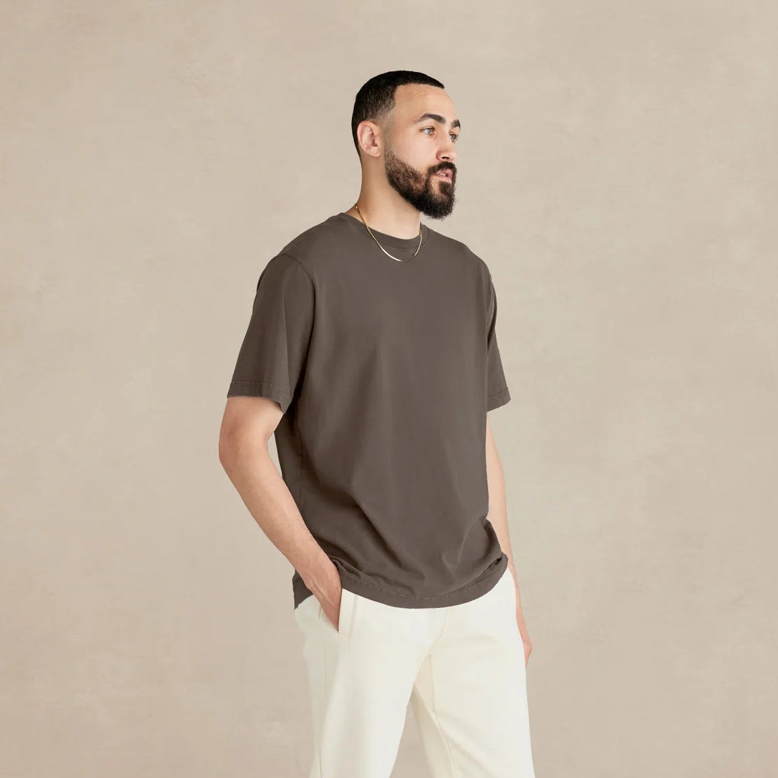 Man wearing a brown t-shirt and white pants standing against a beige background