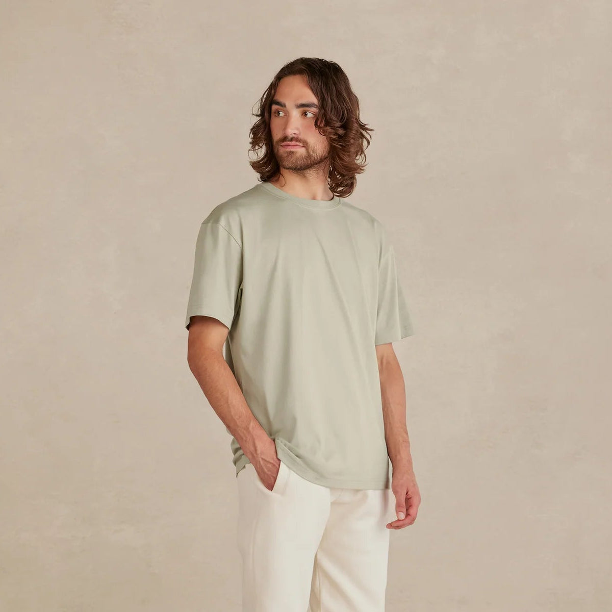 Man wearing a light green t-shirt, white pants, and black sneakers against a beige background