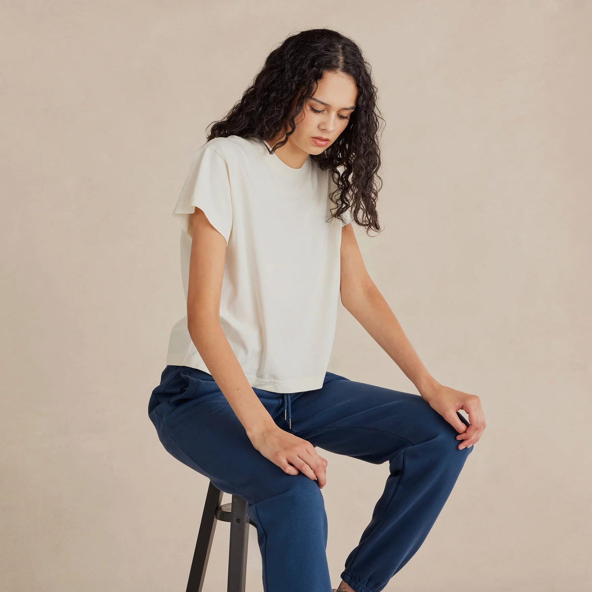 Woman sitting on a stool wearing a white t-shirt and blue jeans against a beige background